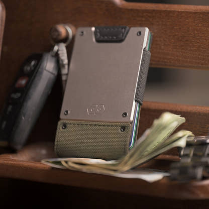 PROOF Operator wallet standing on a wooden surface beside a car key fob and loose cash, satin gunmetal plate above olive drab nylon