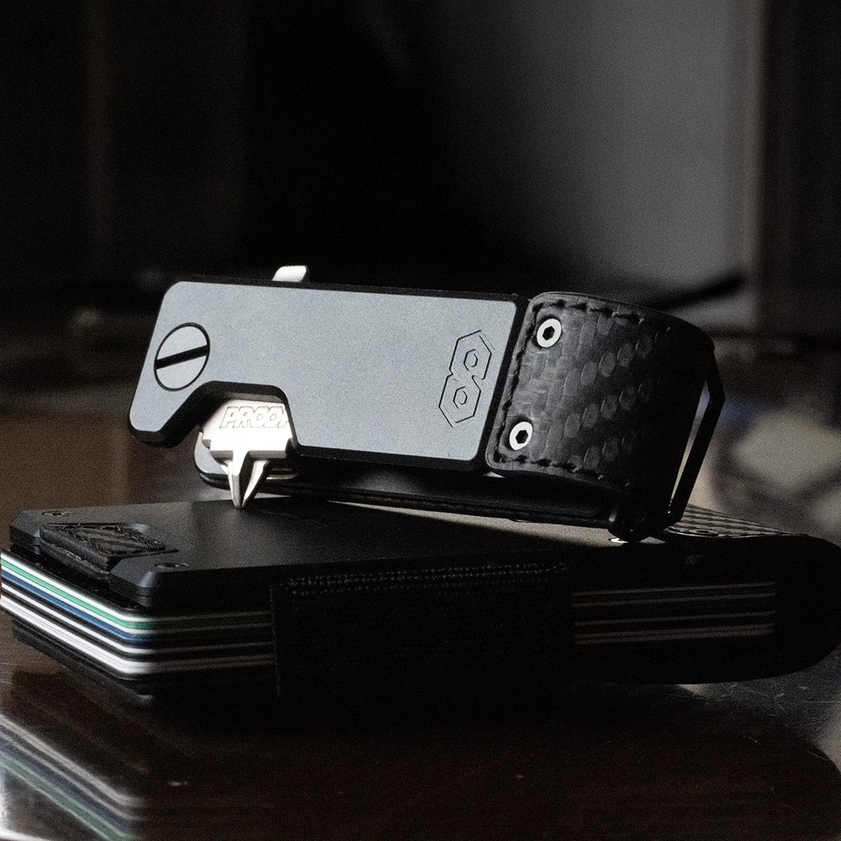 PROOF Oracle key holder in low light with a single key swung out, sitting on a PROOF wallet with card edges visible