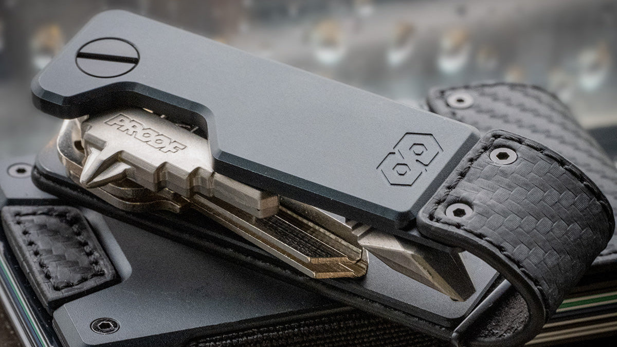 Close-up of the PROOF Oracle key holder with its keys splayed open, carbon fiber weave and gunmetal plate sharp against a blurred background