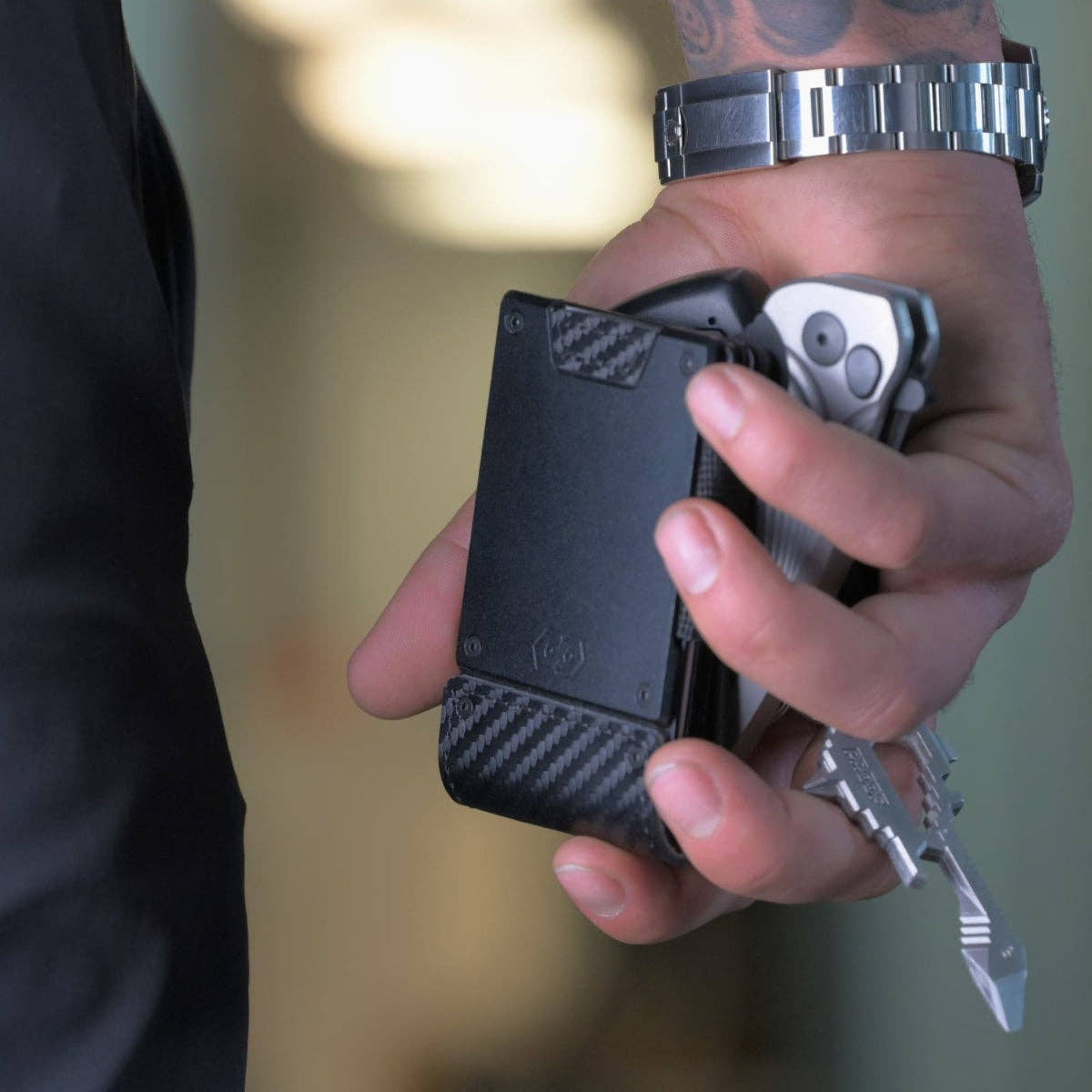 Man gripping the PROOF Oracle carbon fiber wallet together with a phone and keys in one hand, gunmetal plate and carbon weave in view