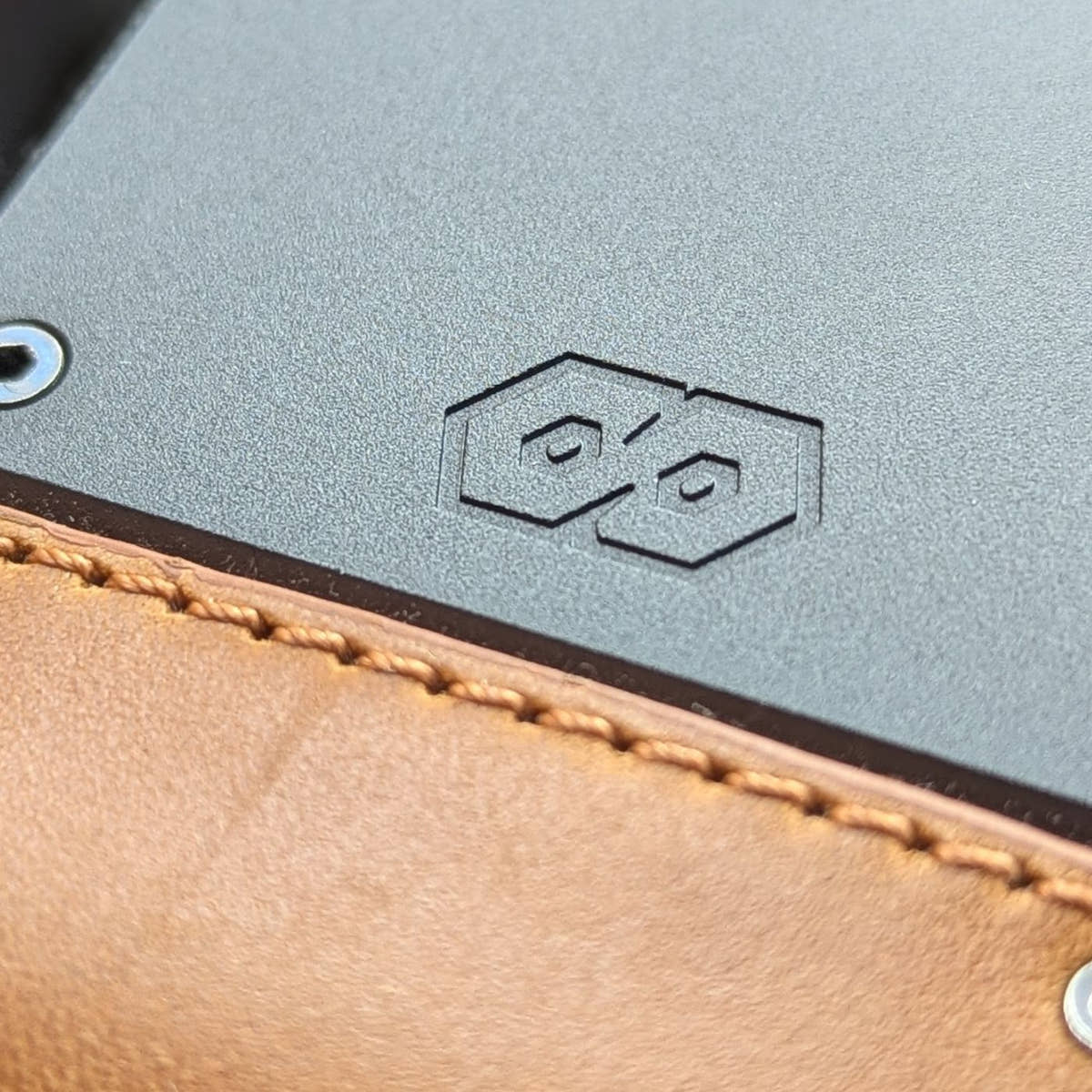Close-up of the PROOF Executive's gunmetal aluminum plate with embossed hexagon logo above tobacco leather